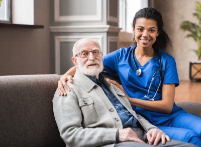Front view portrait of caring afro nurse taking care of an elderly caucasian man grandfather grandpa indoors. Happy female doctor consulting old man in clinic.