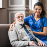 Front view portrait of caring afro nurse taking care of an elderly caucasian man grandfather grandpa indoors. Happy female doctor consulting old man in clinic.